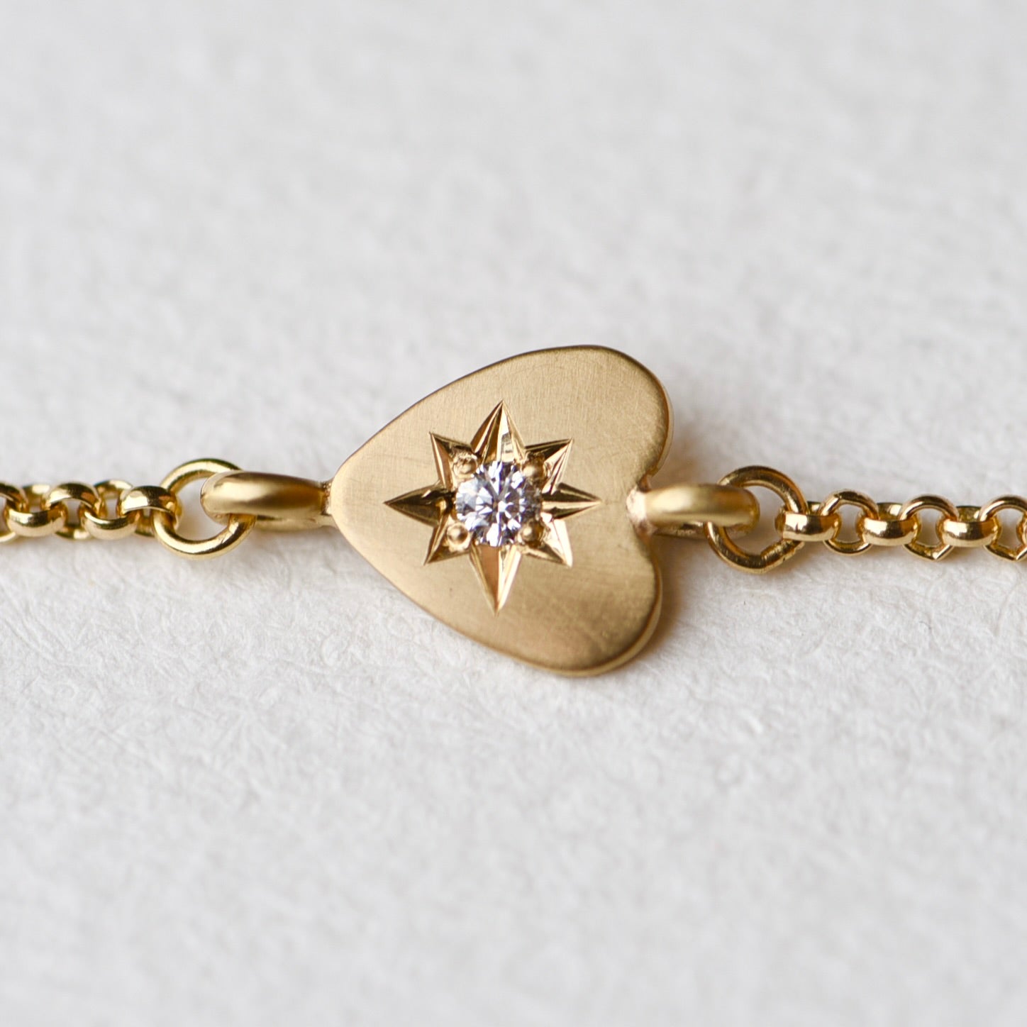 Close-up of the 18k yellow gold bracelet featuring a hand-engraved heart charm with a 2.5mm responsibly sourced diamond set in a star pattern, displayed on a neutral background.