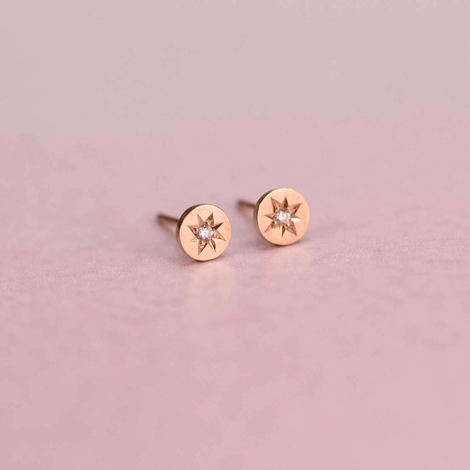 Close-up of Celestial Diamond Studs on a White Background – A pair of small, delicate diamond studs set in gold with a starburst design. Each stud features a sparkling round diamond in the center, surrounded by fine, textured details that give a celestial look.