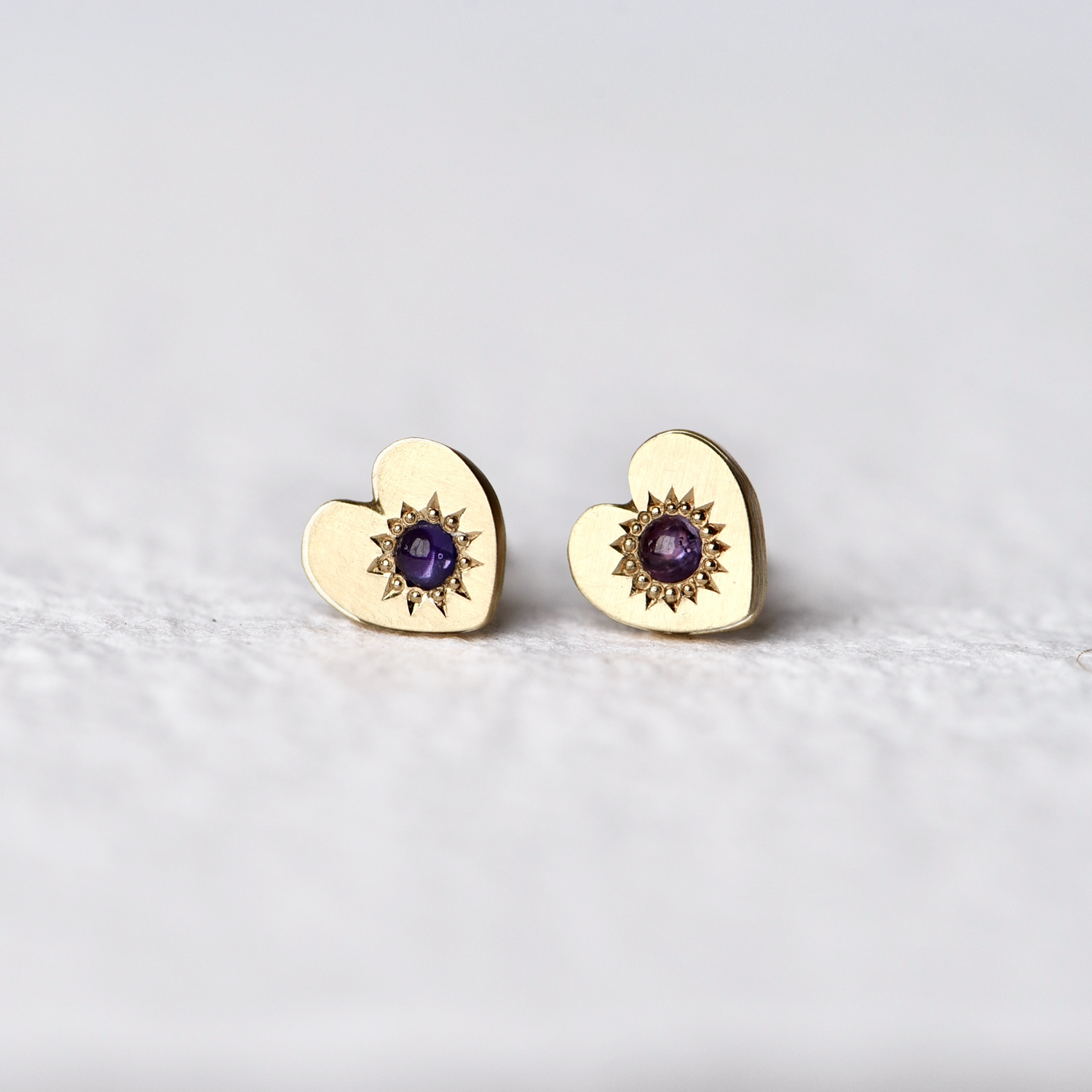 Close-up of Misfit 14k Gold Studs on a white background: A detailed view of the Misfit 14k gold heart-shaped studs, each featuring a color-treated purple sapphire cabochon, showcasing their unique handcrafted design and matte finish.