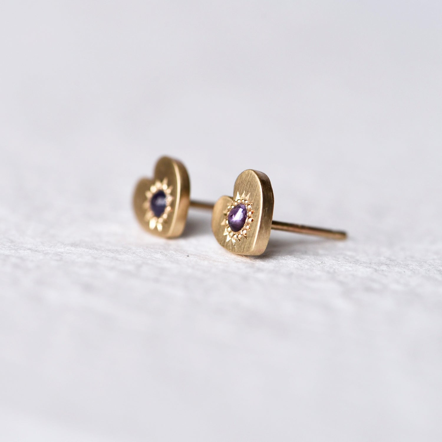 Side angle of Misfit 14k Gold Studs on a white background: A detailed view of the Misfit 14k gold heart-shaped studs, each featuring a color-treated purple sapphire cabochon, showcasing their unique handcrafted design and matte finish.