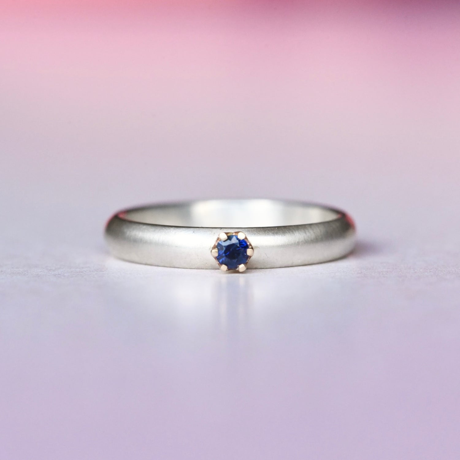 Silver ring with a 14k yellow gold prong set blue sapphire on a gradient pink and purple background
