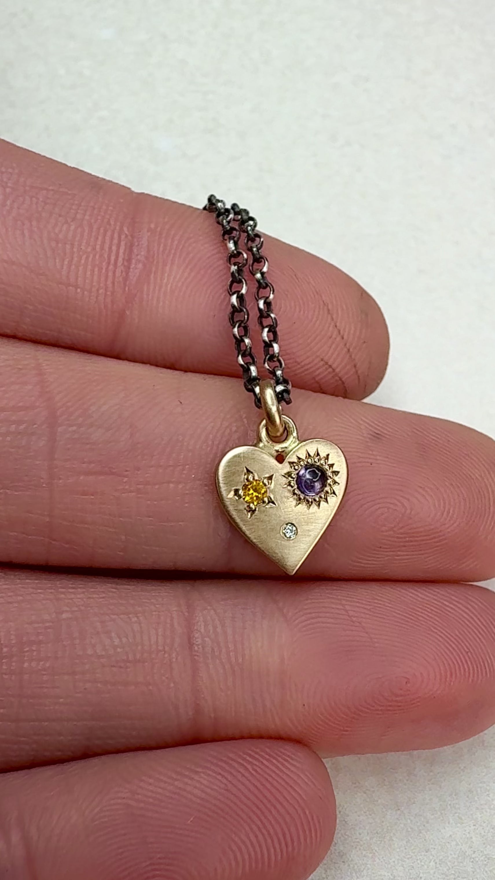 Video of the HELL YES 14k Gold Sapphire and Diamond Misfit Heart Necklace rotating under natural light, showing the yellow and purple sapphires, diamond sparkle, and hand-engraved “HELL YES” on the back. Handmade by Corkie Bolton Jewelry.