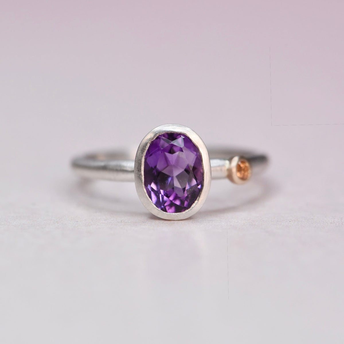 Front view of a sterling silver ring with a bezel-set oval amethyst and a small orange sapphire accent in a 14k gold bezel on the band