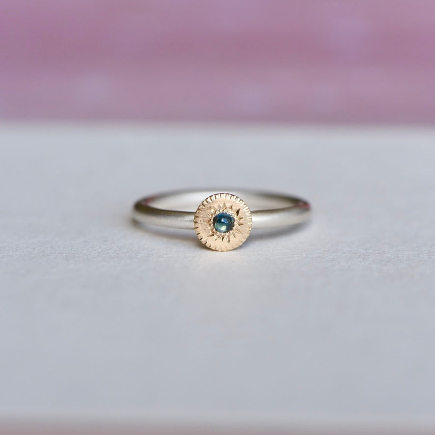 Mixed-metal ring featuring a hand-engraved 14k gold disc with a blue sapphire set on a satin-finish sterling silver band, shown from the front on a soft background.
