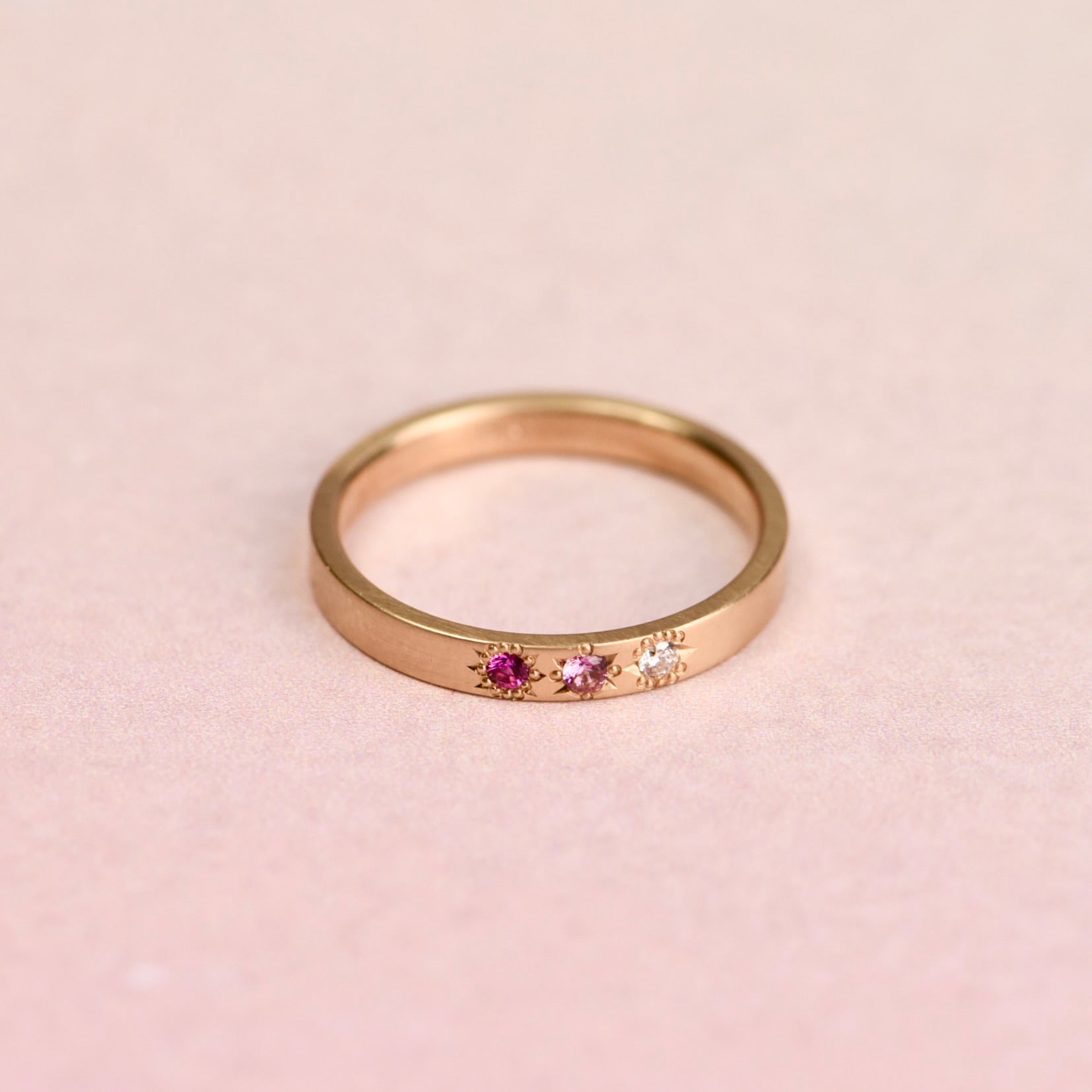 Top view of a 14k yellow gold ring featuring two pink sapphires and a misfit diamond in hand-engraved star settings, designed by Corkie Bolton Jewelry.