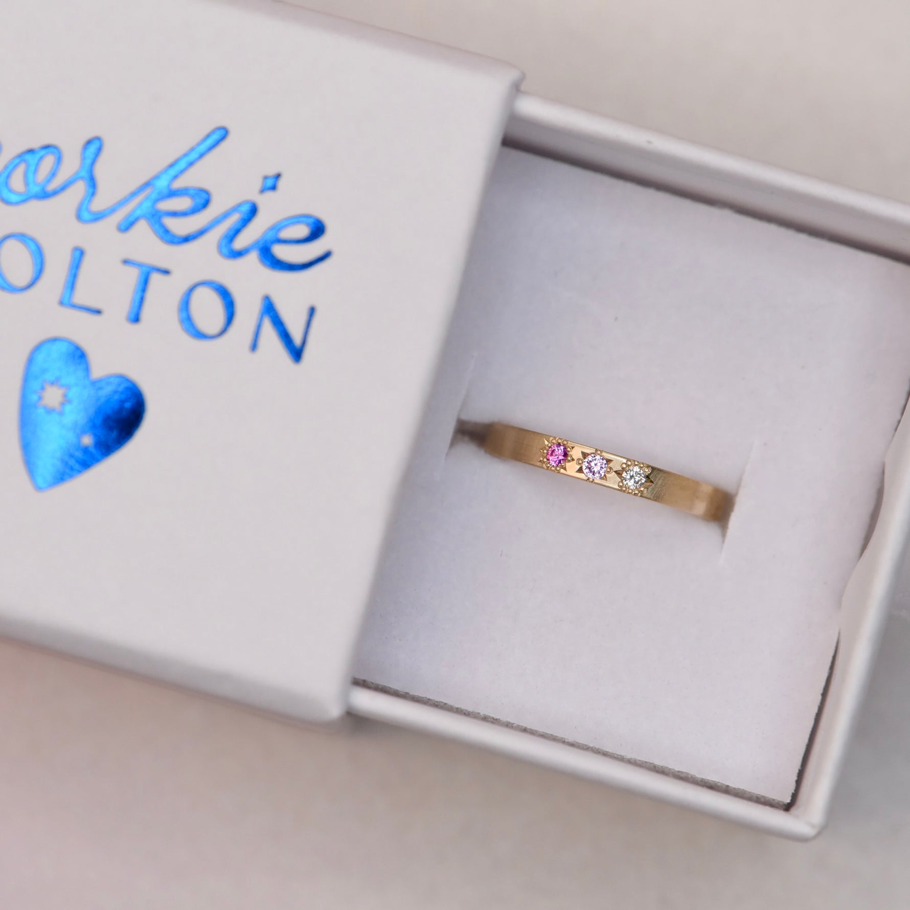 14k gold pink sapphire and diamond ring resting inside a white jewelry box with “Corkie Bolton” in neon blue hot foil, showcasing elegant packaging and craftsmanship.