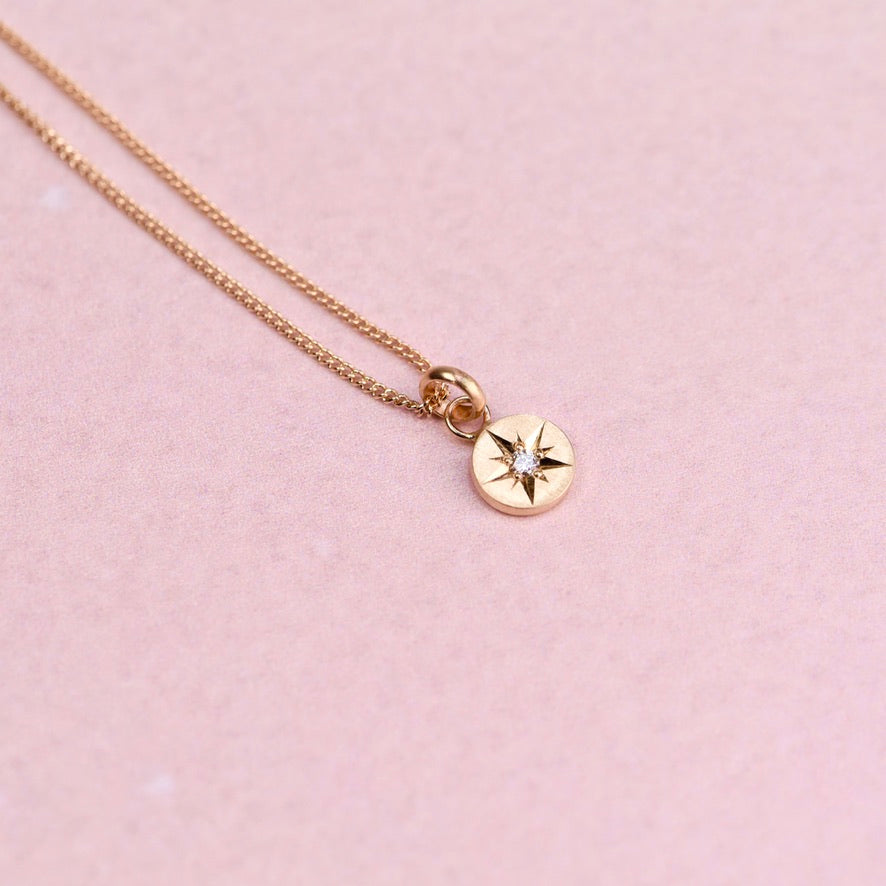 Flatlay of a 14k gold necklace featuring a hand-engraved star-set diamond charm on a curb chain against a soft pink background