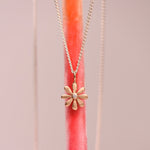 Matte 14k yellow gold flower charm pendant with flush set diamond center hanging on a sterling silver curb chain against a pink display stand