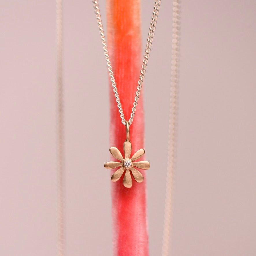 Matte 14k yellow gold flower charm pendant with flush set diamond center hanging on a sterling silver curb chain against a pink display stand