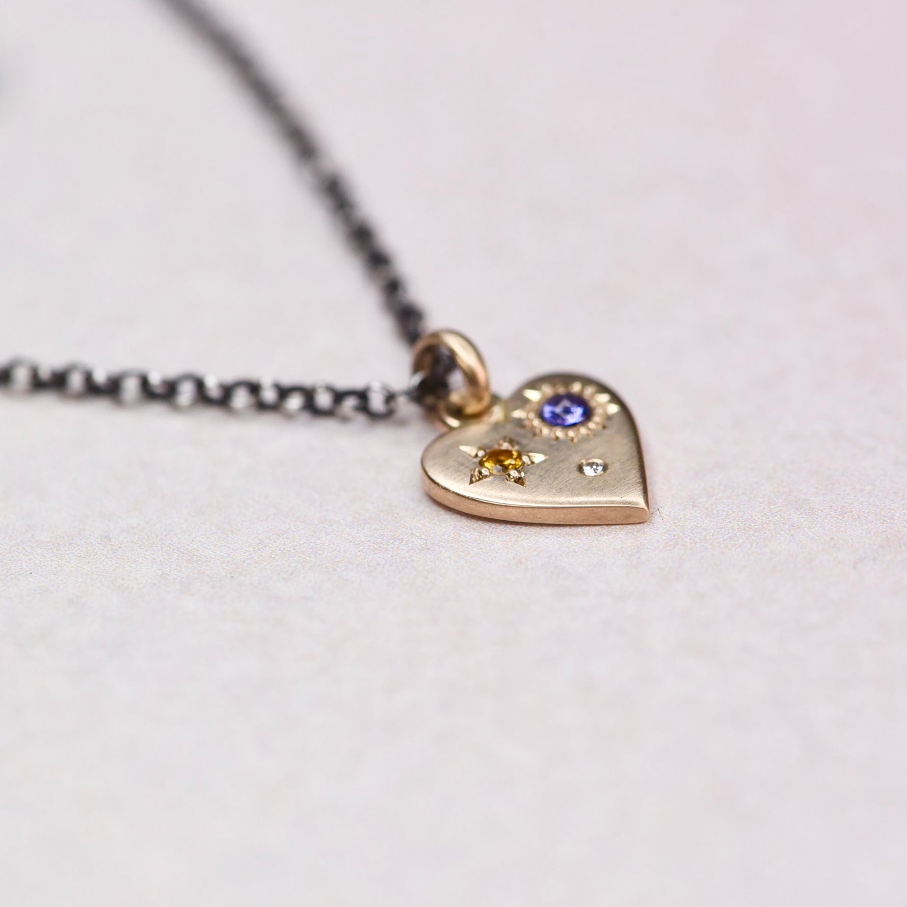 Detail view of the 14k gold Misfit Heart charm featuring a yellow faceted sapphire, purple cabochon sapphire, and diamond, showing fine engraving and satin finish by Corkie Bolton Jewelry.