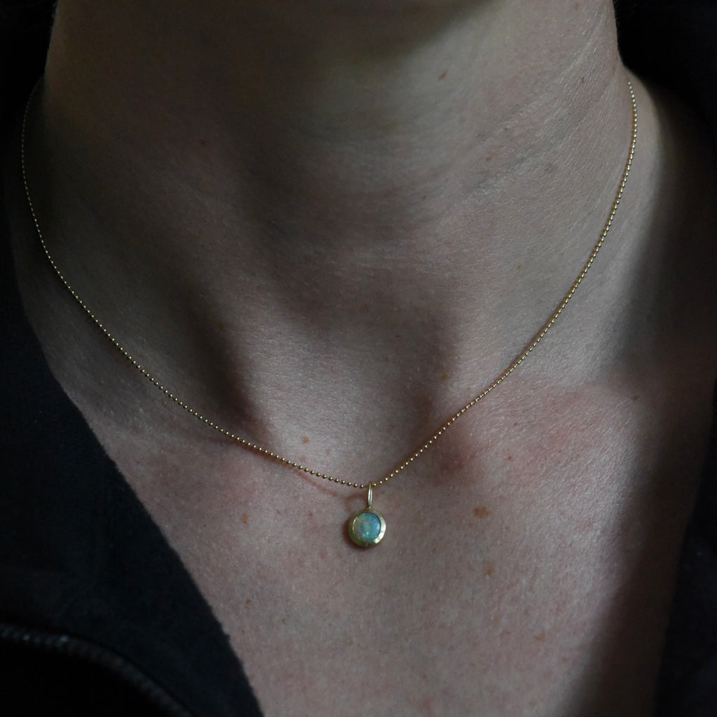 A gorgeous opal charm set in 14k yellow gold on a 14k yellow gold ball chain being worn.