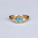 A black opal ring with blue and green flashes, set in a 14k yellow gold tapered band with a satin finish, on a white background.