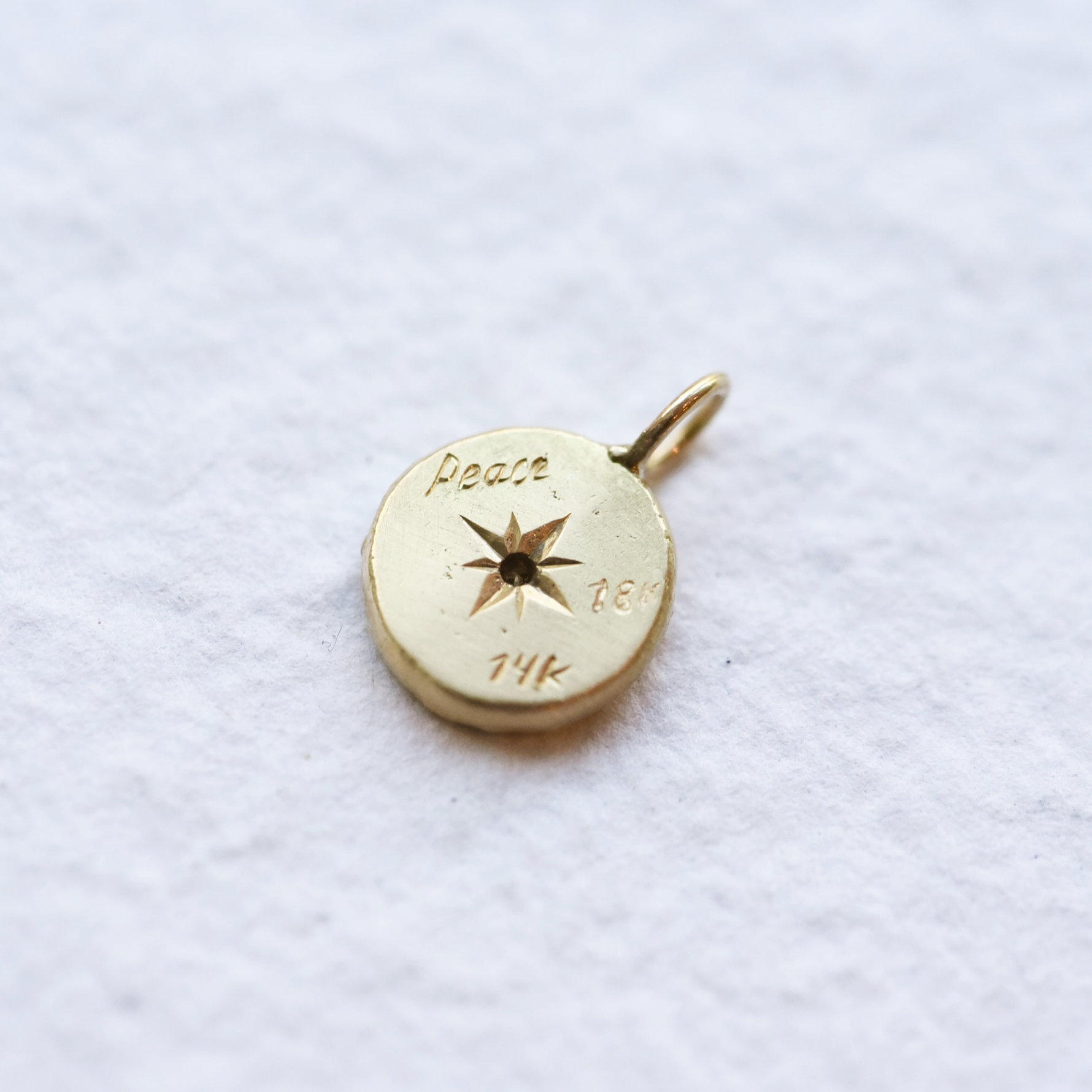 Backside of Detail shot of the hand-engraved design on the Reborn 18k Gold Charm, capturing the fine artistry and texture. Showing an engraved 'Peace' '14k' '18k'.