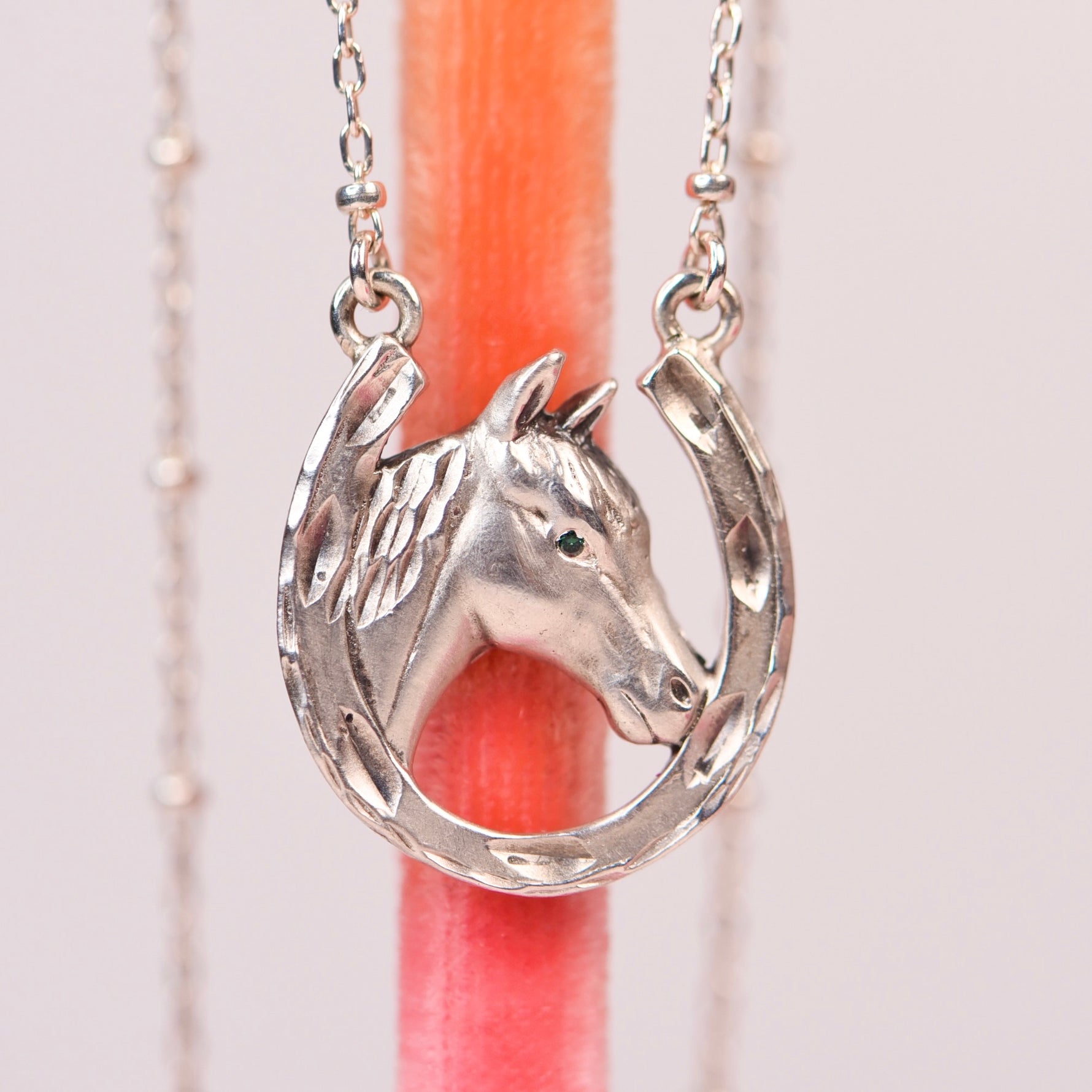 A silver horseshoe pendant necklace featuring a detailed horse head and horseshoe design, hanging against a soft pink background with a blurred coral-colored vertical prop.