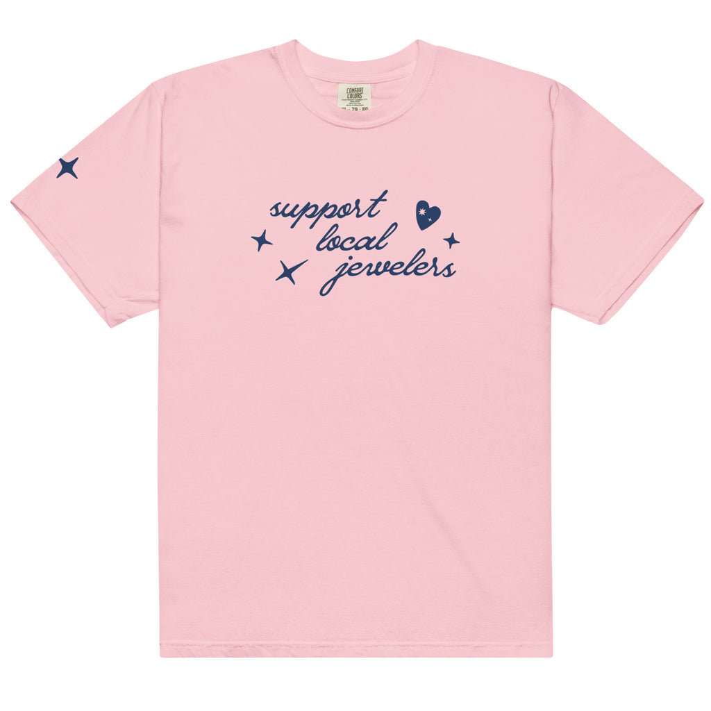 Light pink, short-sleeve T-shirt with a navy blue design on the front. The design features the phrase "support local jewelers" in cursive font, surrounded by small star-like shapes and a heart with a sparkle inside. There is also a small star detail on the left sleeve.