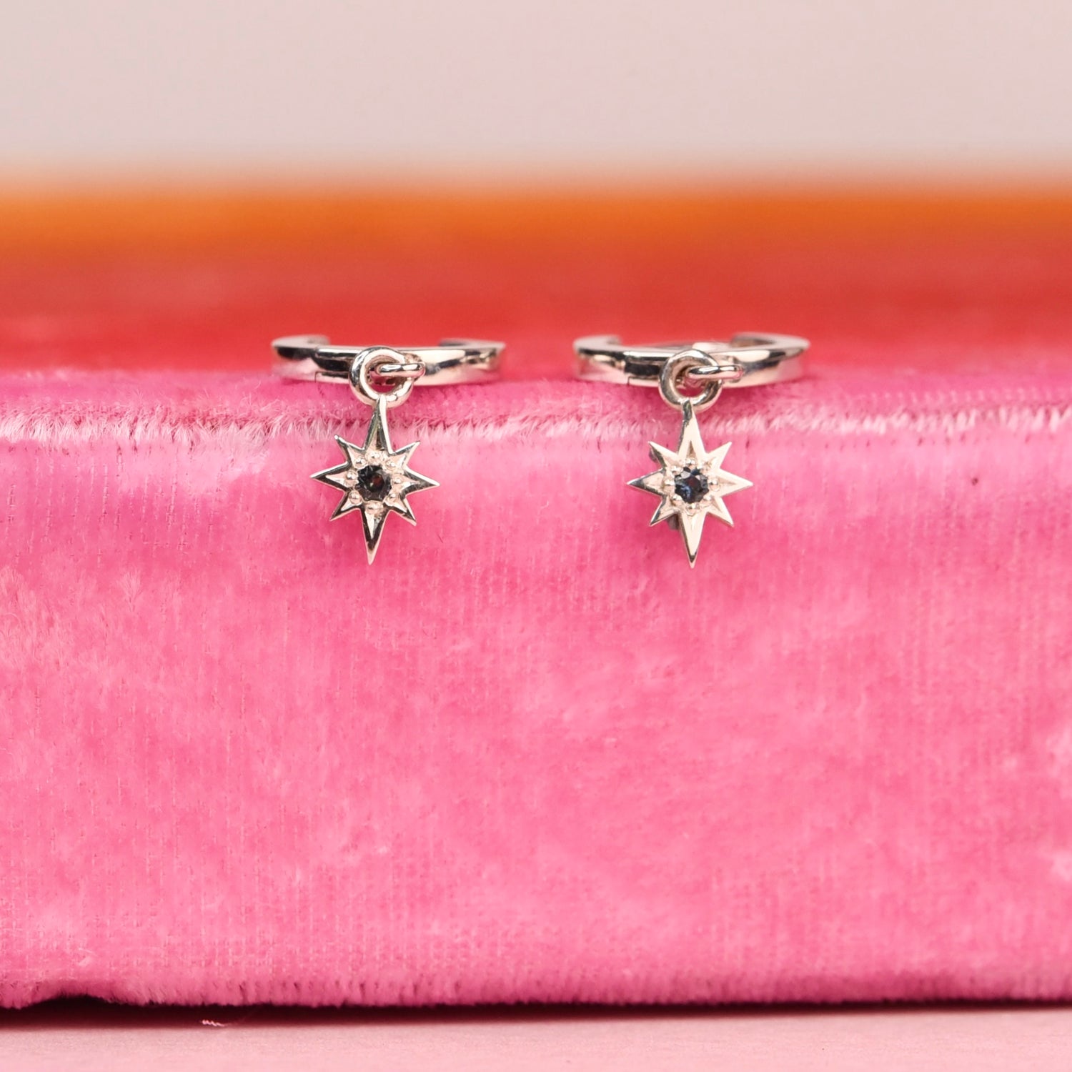 Top view of sterling silver star dangle huggie hoop earrings with blue sapphires on pink velvet background