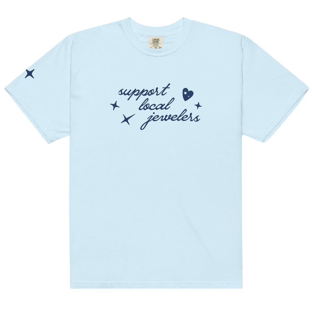 Light blue, short-sleeve T-shirt with a navy blue design on the front. The design features the phrase "support local jewelers" in cursive font, surrounded by small star-like shapes and a heart with a sparkle inside. A small star detail is also present on the left sleeve.