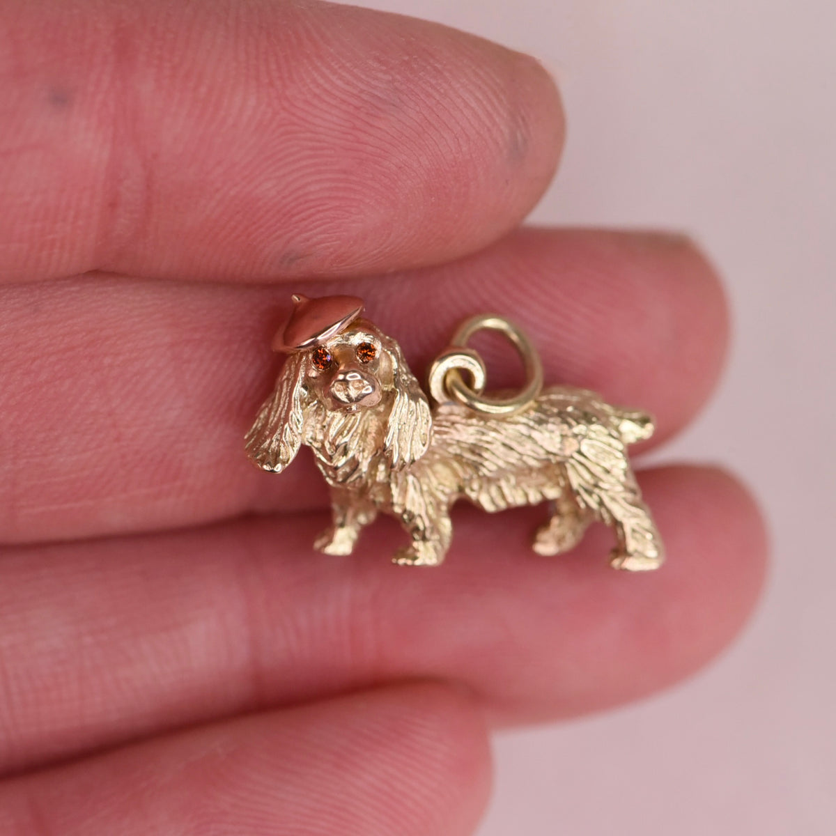 Vintage gold cocker spaniel dog charm with a rose gold beret and orange diamond eyes held on a fingertip to show its size