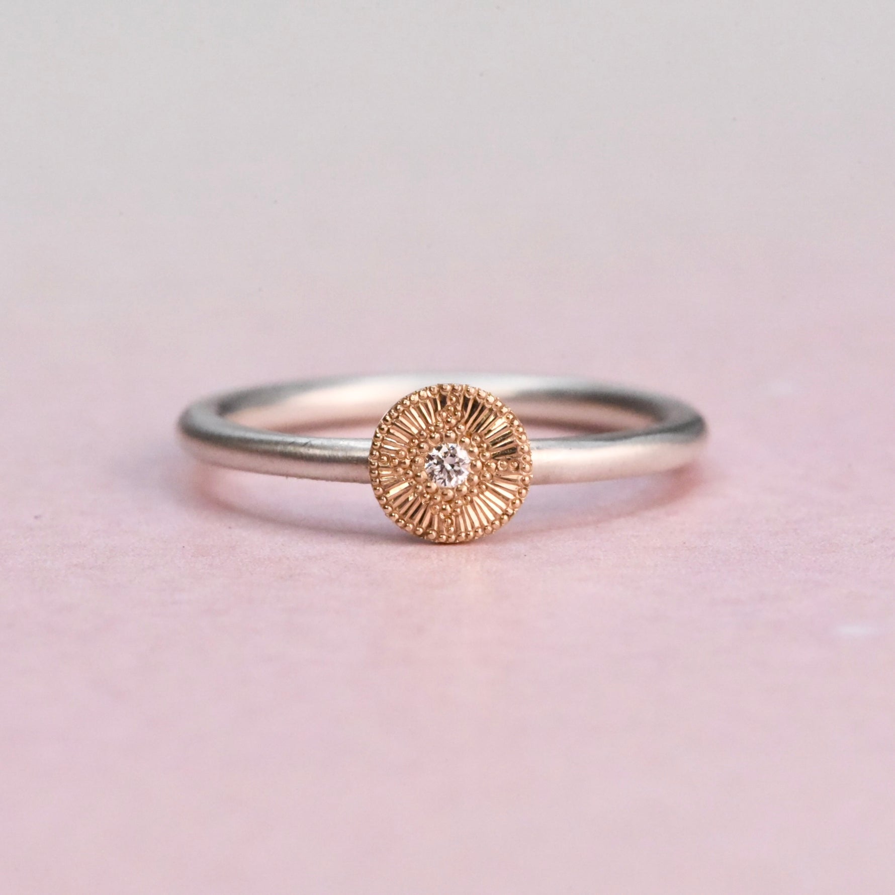 Silver ring with a gold circular design featuring a diamond on a light pink background