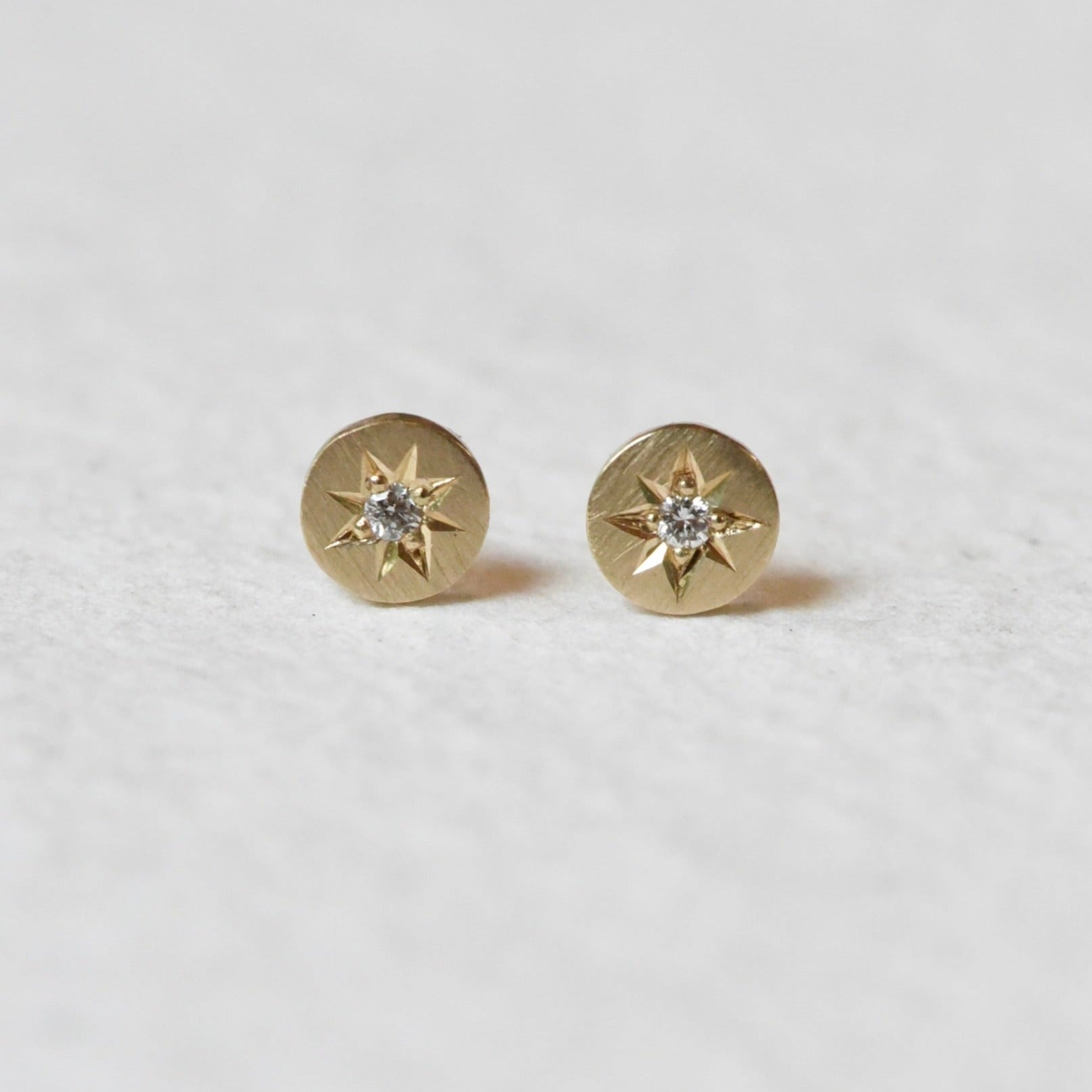 Close-up of Celestial Diamond Studs on a White Background – A pair of small, delicate diamond studs set in gold with a starburst design. Each stud features a sparkling round diamond in the center, surrounded by fine, textured details that give a celestial look.