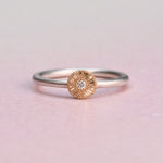 Silver ring with a gold circular design featuring a diamond on a light pink background