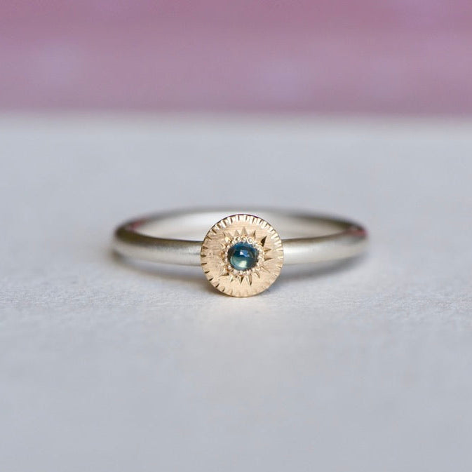 Mixed-metal ring featuring a hand-engraved 14k gold disc with a blue sapphire set on a satin-finish sterling silver band, shown from the front on a soft background.