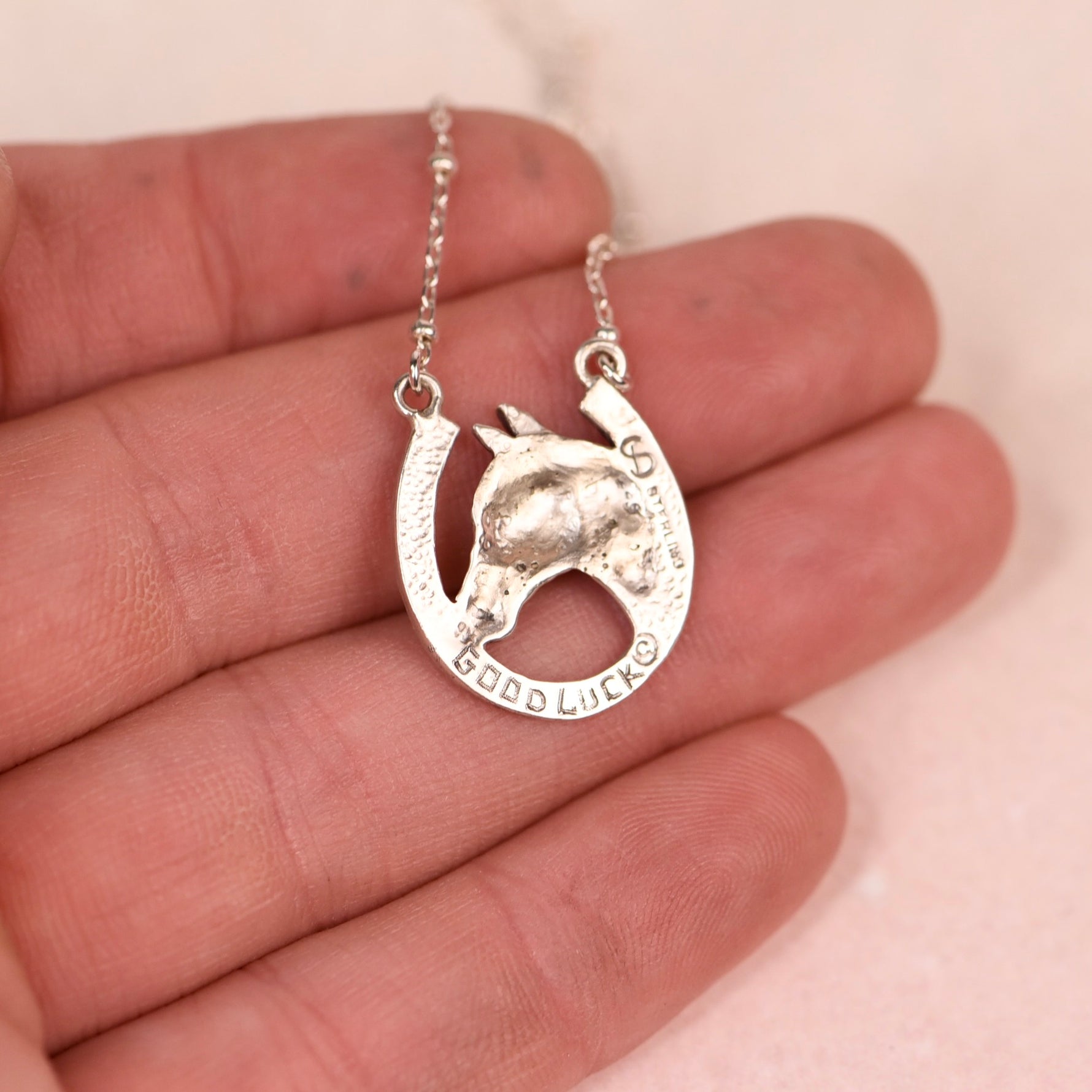 Backside of a sterling silver necklace with a horseshoe pendant featuring a horse's head and the words 'GOOD LUCK' engraved, held in a person's hand against a soft pink background.