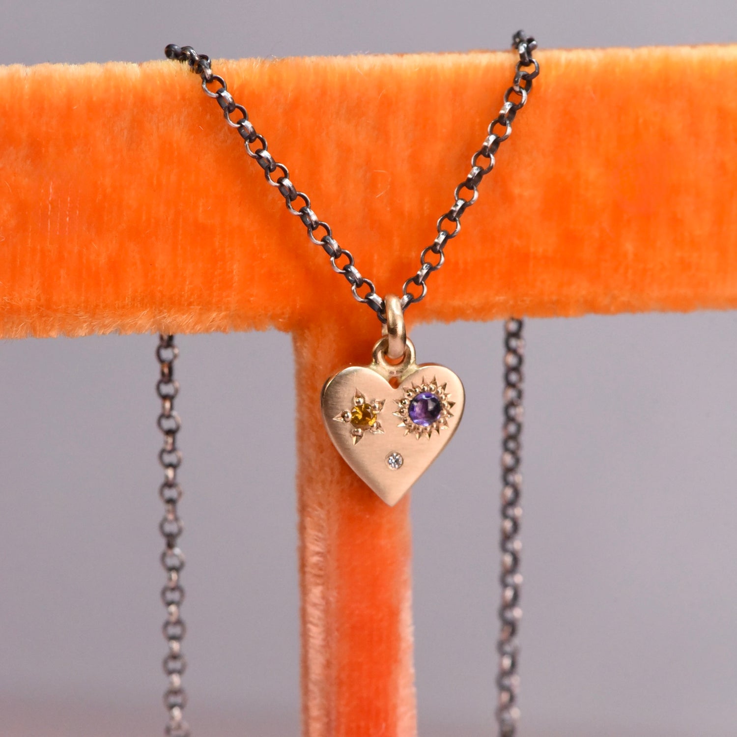 Front view of the handmade 14k yellow gold Misfit Heart necklace featuring a yellow faceted sapphire, a purple cabochon sapphire, and a diamond on an oxidized silver chain by Corkie Bolton Jewelry.