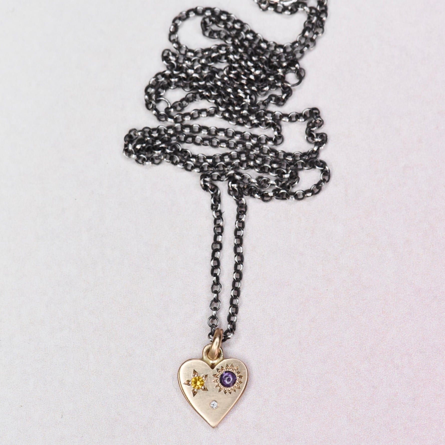 Full view of the handmade HELL YES Misfit Heart Necklace by Corkie Bolton Jewelry, featuring a 14k yellow gold heart pendant with yellow and purple sapphires and a diamond on an oxidized sterling silver rolo chain.