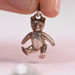 A close-up of a small metallic teddy bear charm being held by fingers against a soft pink background. 