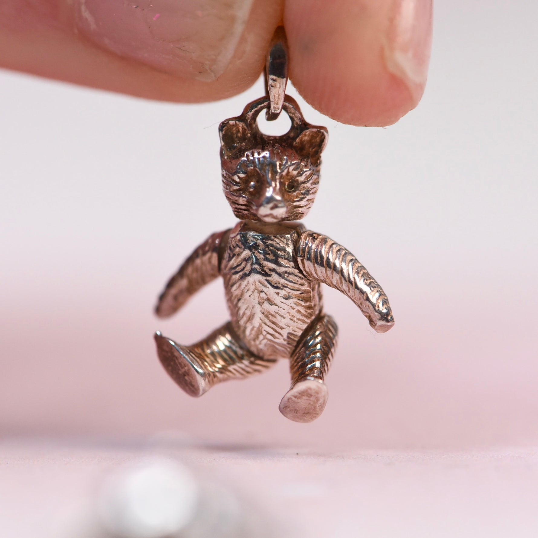 A close-up of a small metallic teddy bear charm being held by fingers against a soft pink background. 