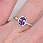 Sterling silver oval amethyst ring worn on a finger, showing the orange sapphire accent in a 14k gold bezel on the side of the band