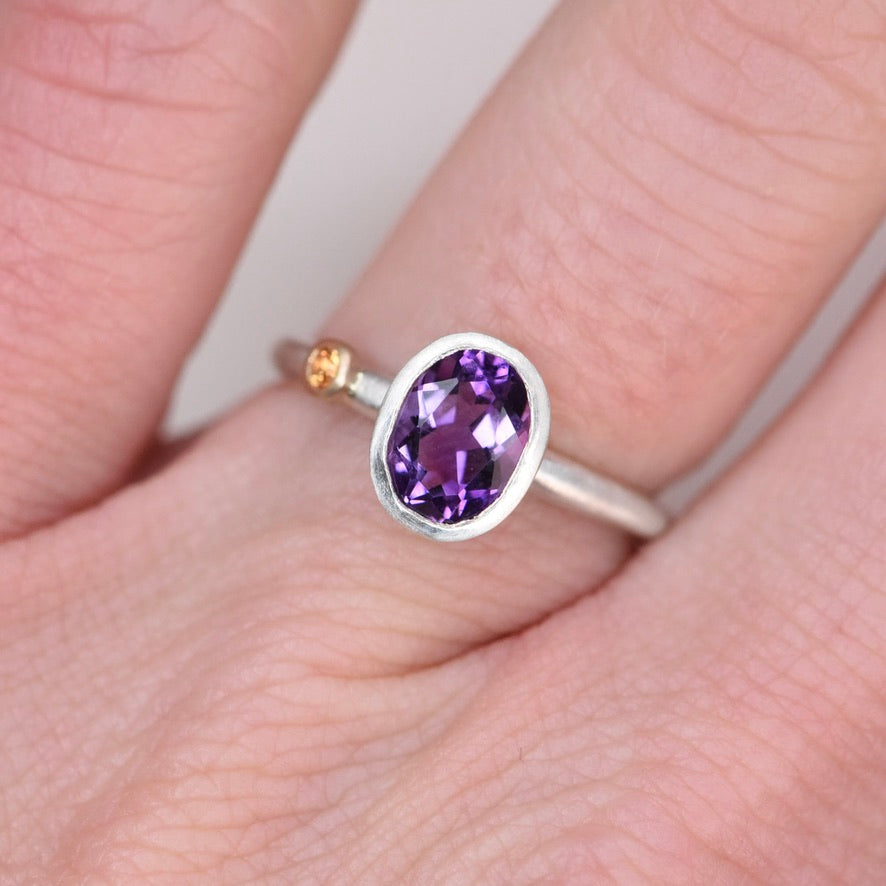 Sterling silver oval amethyst ring worn on a finger, showing the orange sapphire accent in a 14k gold bezel on the side of the band