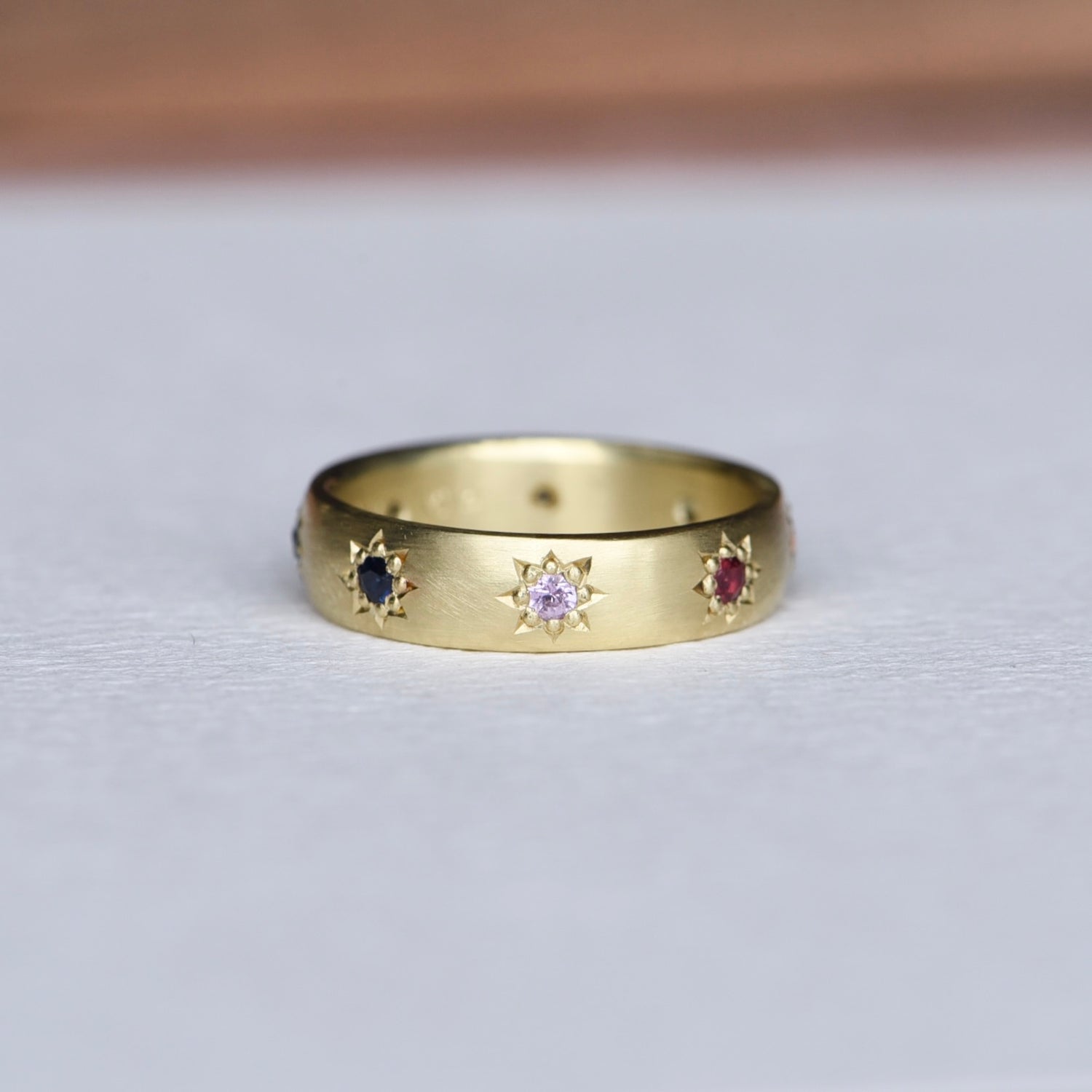 Rainbow Love 18k Gold Ring - Size 7. A wide satin finish, buttery yellow 18k gold band featuring hand-engraved star settings adorned with a rainbow of sapphires and diamonds. The luxurious texture and vibrant gemstones create a bold, elegant statement.