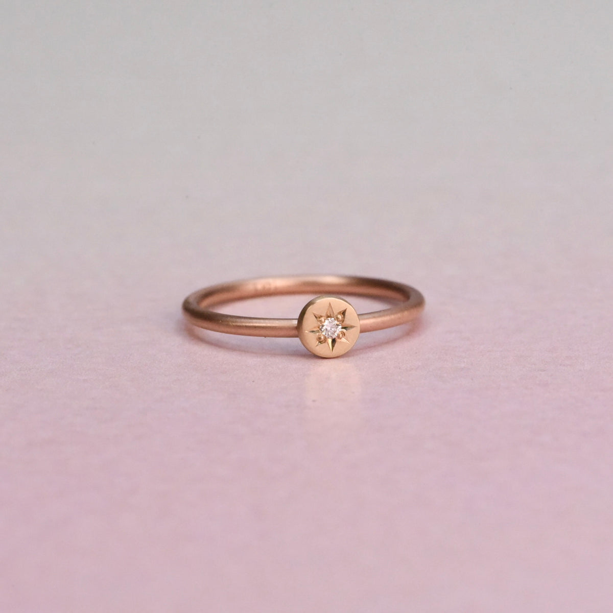 A delicate rose gold ring with a small star-shaped diamond set on a 14k yellow disc with a white diamond. on a pionk background