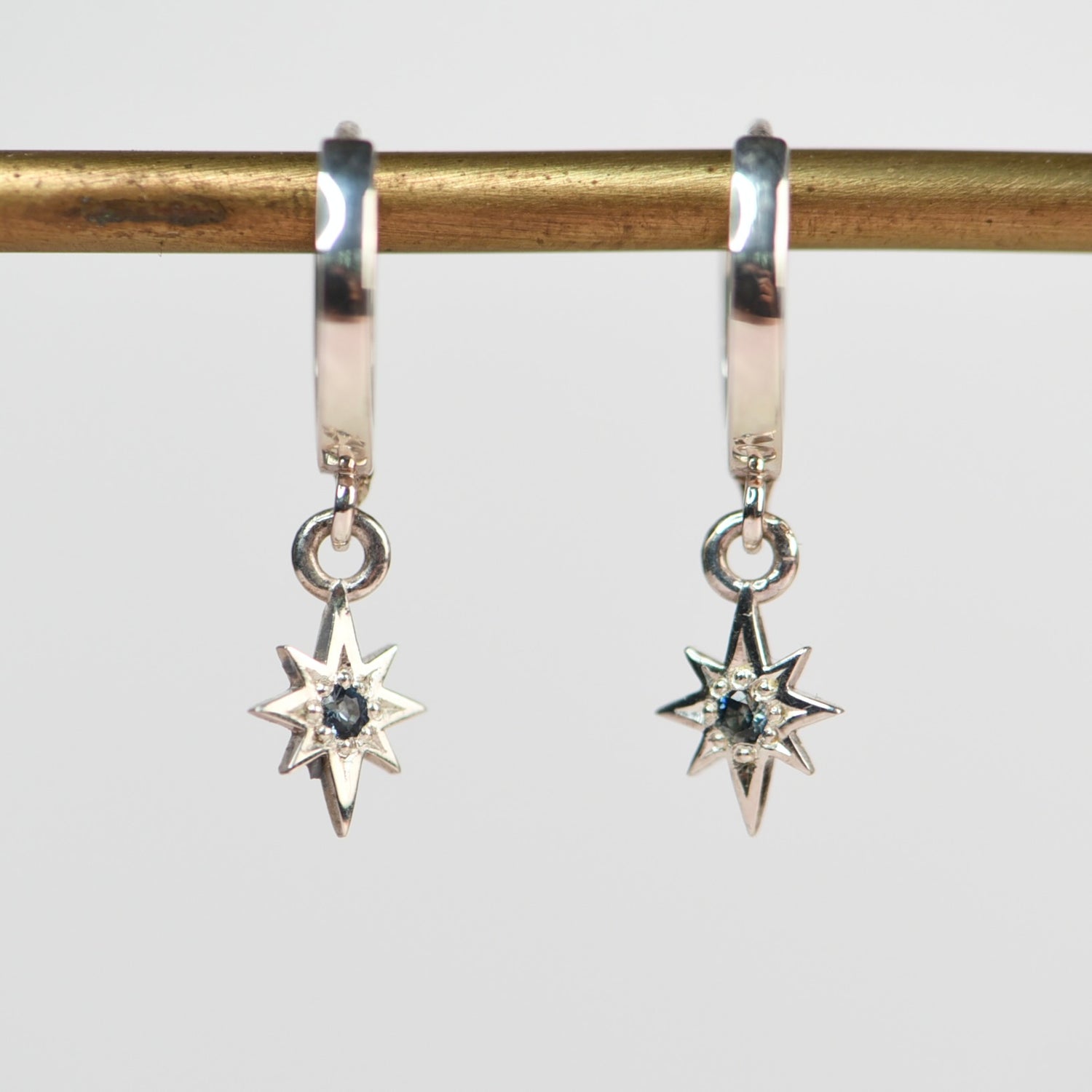 Pair of sterling silver dangle huggie hoop earrings with engraved star charms and blue sapphire centers hanging from a brass rod

