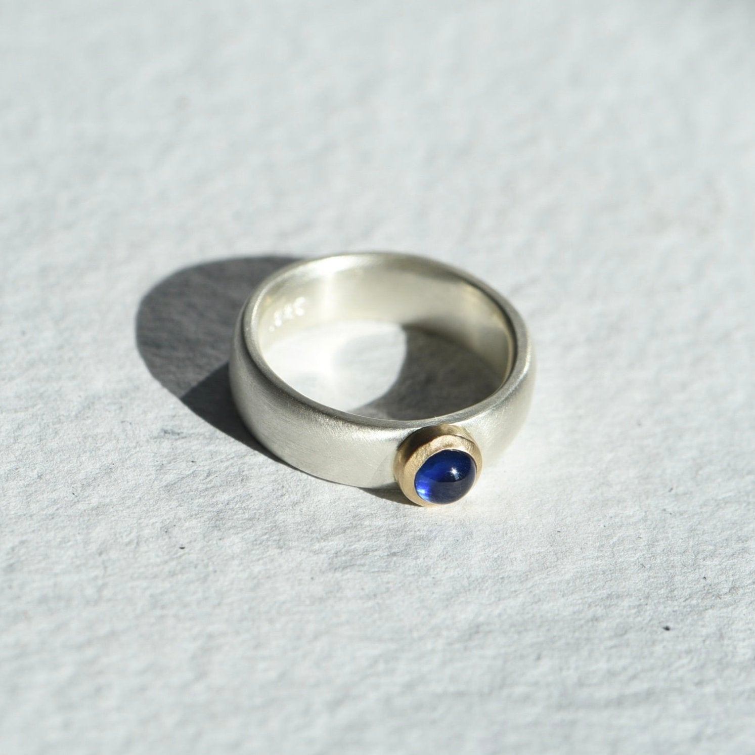 Strength Sapphire Mixed Ring on a white background, featuring a deep blue sapphire set in a 14k gold bezel with a satin finish sterling silver band. Created by Corkie Bolton.