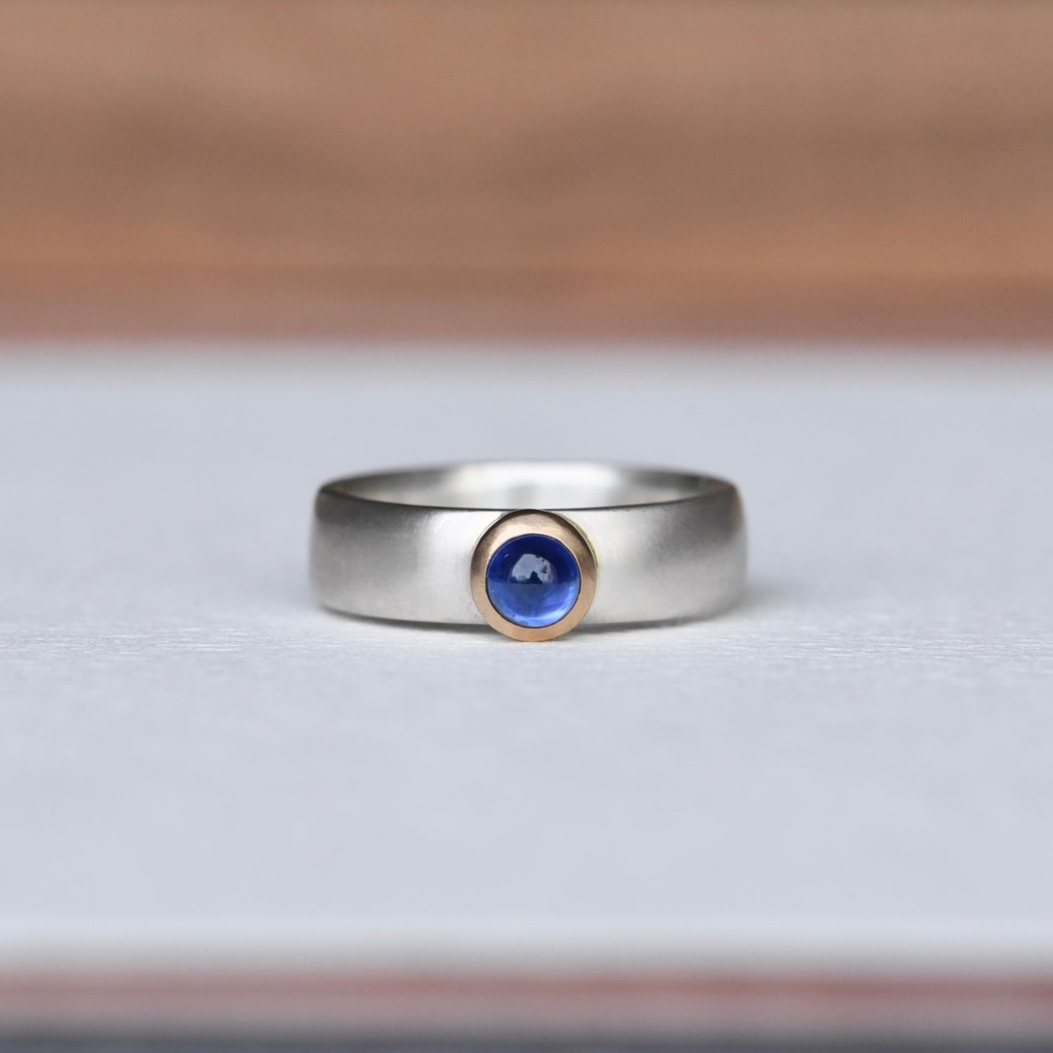 Strength Sapphire Mixed Ring on a white background, featuring a deep blue sapphire set in a 14k gold bezel with a satin finish sterling silver band. Created by Corkie Bolton.