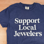 Navy blue t-shirt with 'Support Local Jewelers' text on a wooden surface
