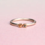 Sterling silver stacker ring with two small sapphires — orange and blue — each set in a 14k gold bezel, on a pink background