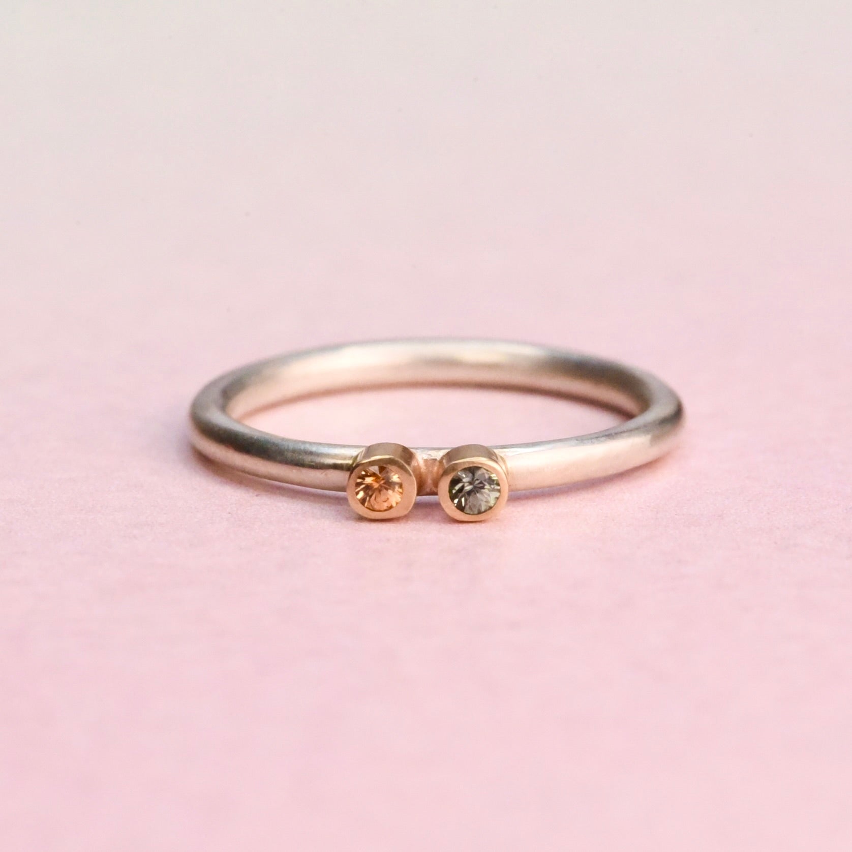 Sterling silver stacker ring with two small sapphires — orange and blue — each set in a 14k gold bezel, on a pink background