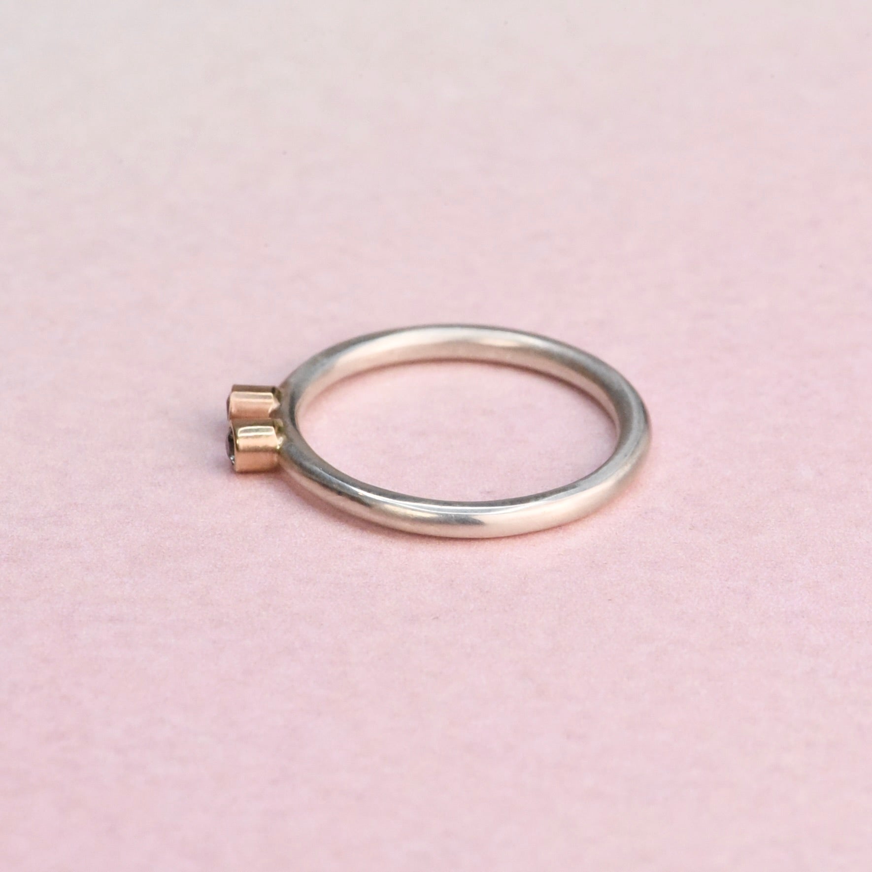 Side view of a slim sterling silver stacker ring with two 14k gold bezels on a pink background