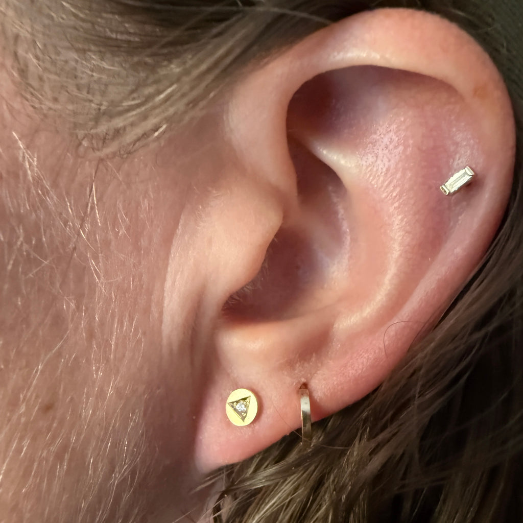 An up-close of an ear with a helix piercing a small gold hoop and the earrings from this product page which are handcrafted 14k gold diamond stud earrings by Corkie Bolton Jewelry featuring unique triangle engravings. These artisan-made earrings highlight delicate, hand-engraved details with a sparkling diamond centerpiece in each stud.