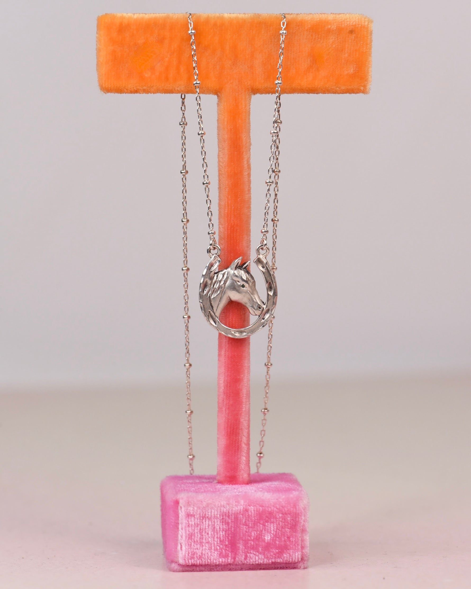 A sterling necklace with a horse ina horseshoe charm displayed on a pink velvet T-stand with an orange top bar against a light gray background.