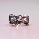 A silver bow-shaped ring with a round purple gemstone in the center, photographed on a soft pink background.