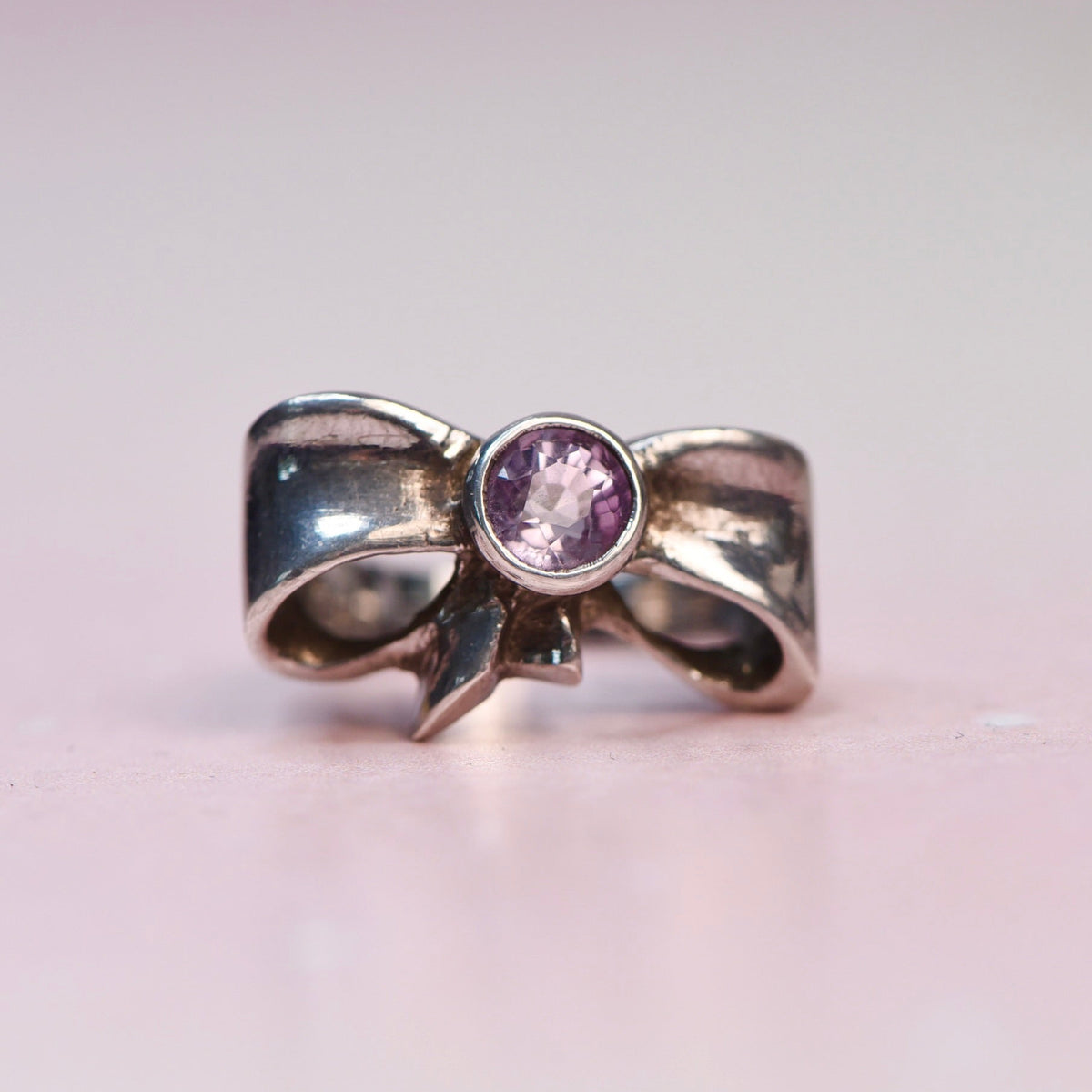 A silver bow-shaped ring with a round purple gemstone in the center, photographed on a soft pink background.