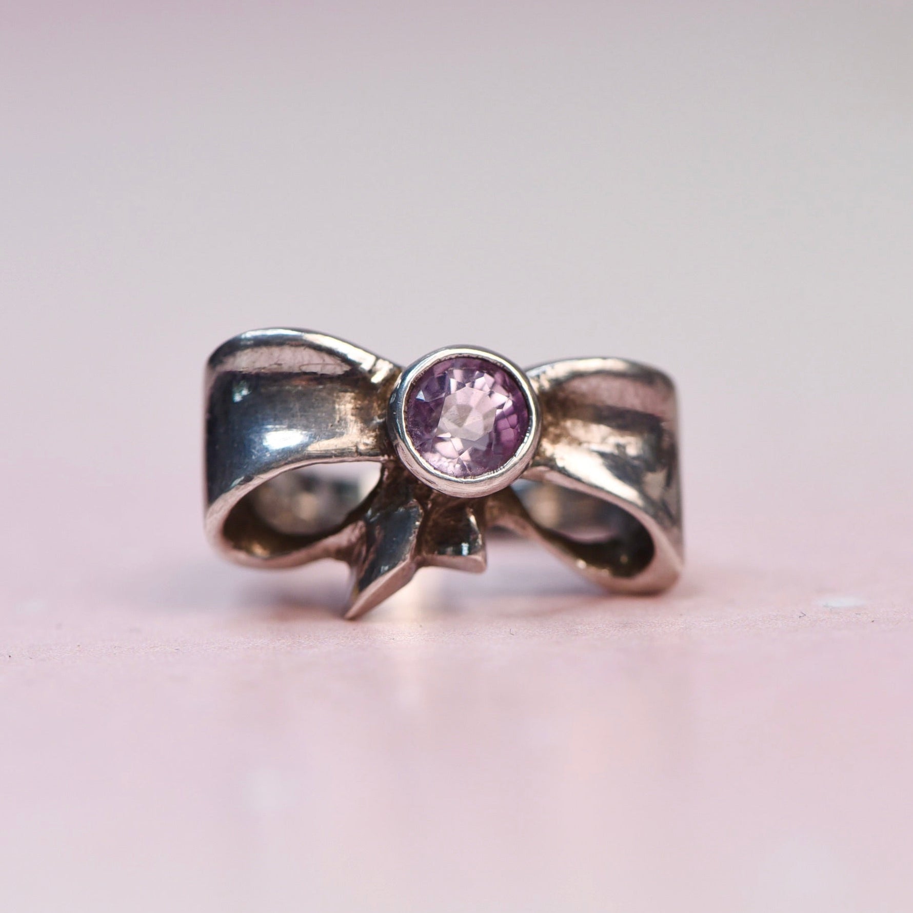 A silver bow-shaped ring with a round purple gemstone in the center, photographed on a soft pink background.
