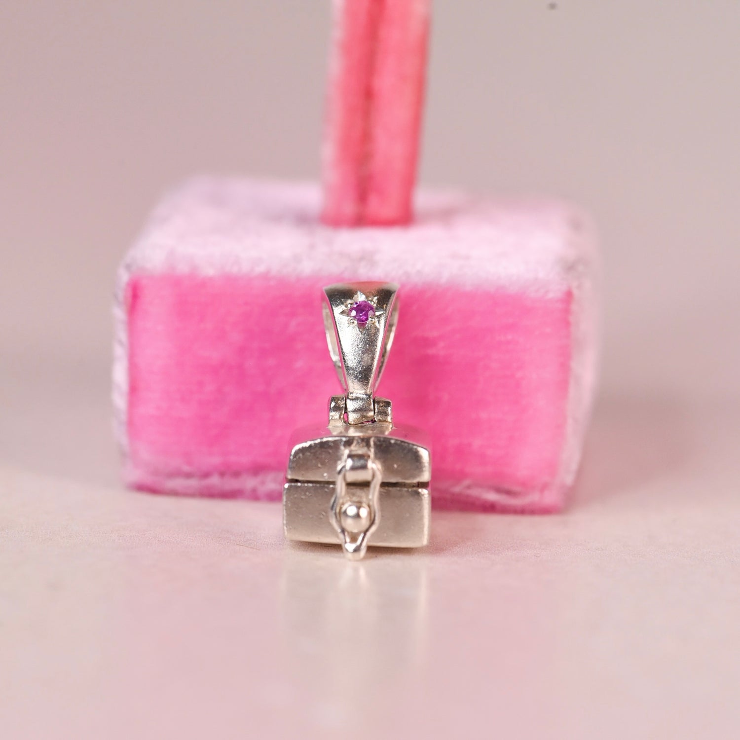 A sterling silver jewelry box pendant with a pink sapphire accent on the bail, displayed in front of a pink velvet jewelry stand.