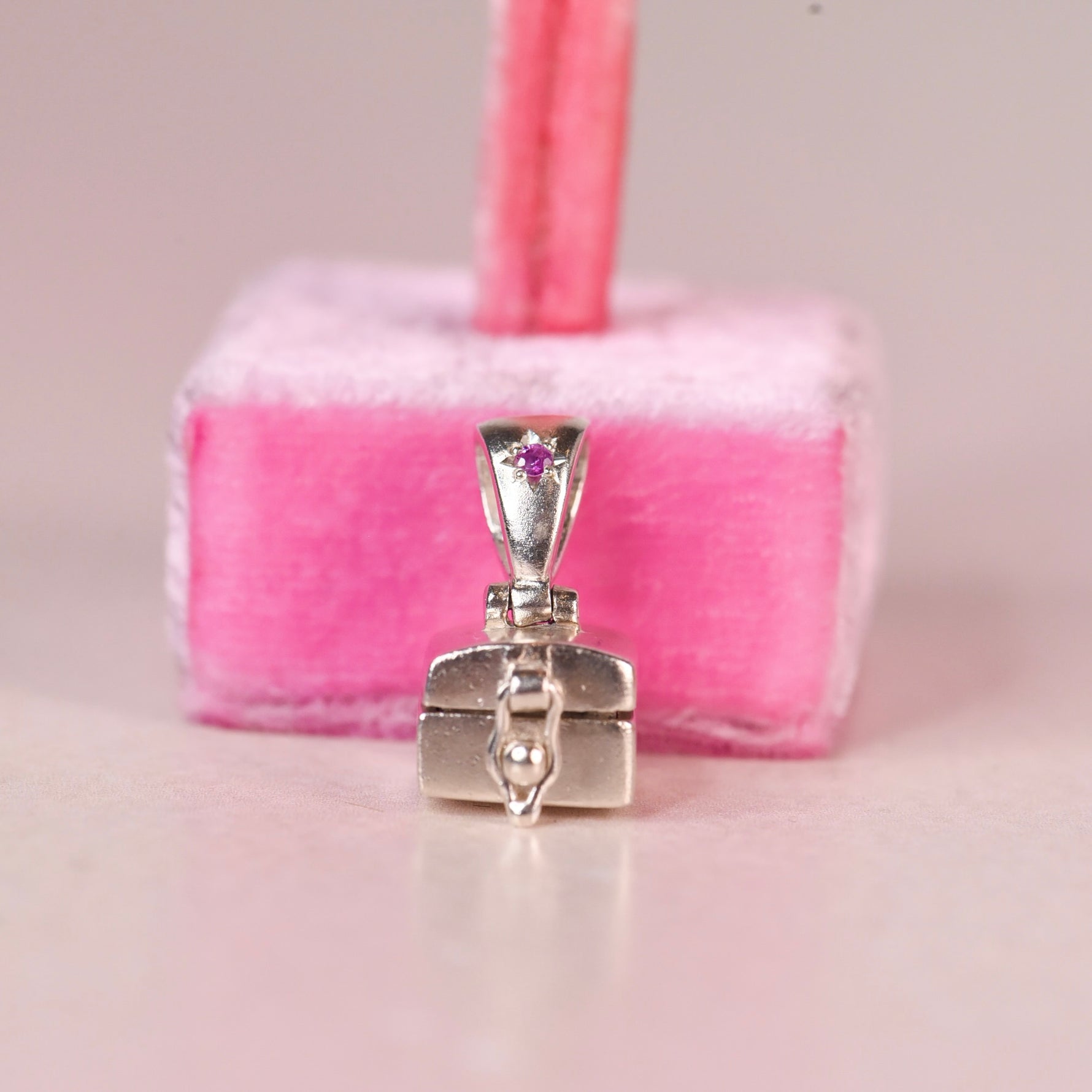 A sterling silver jewelry box pendant with a pink sapphire accent on the bail, displayed in front of a pink velvet jewelry stand.