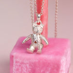 A silver teddy bear pendant necklace with a sorange diamond on belly. displayed on a pink velvet stand against a soft pink background.
